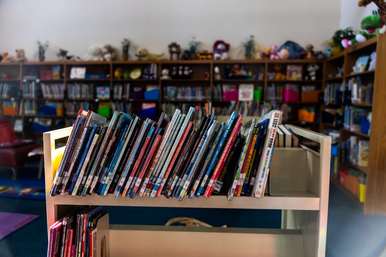 A warm library interior featuring a book cart in focus with colorful children's books on shelves.